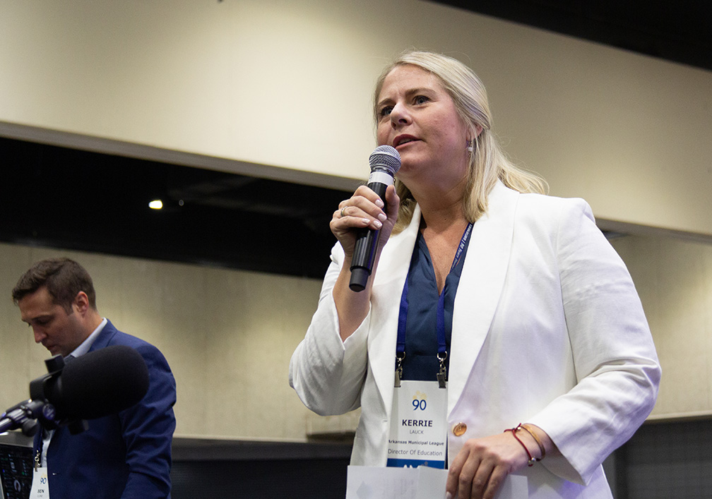 A blonde white woman wearing a white blazer and blue top is speaking into a microphone.