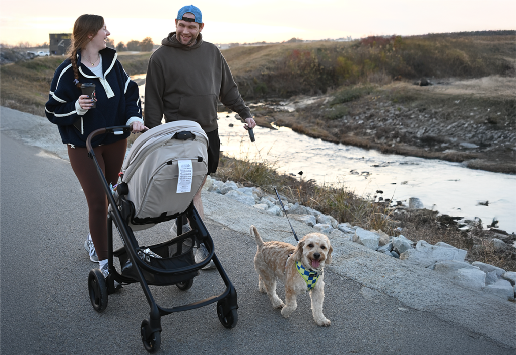 A man and woman walking and talking on a walking trail while pushing a baby stroller. A small terrier in a snazzy checkered harness walks alongside the couple.